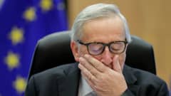 Jean-Claude Juncker silmät kiinni ja käsi suunsa edessä EU-lippu taustallaan.