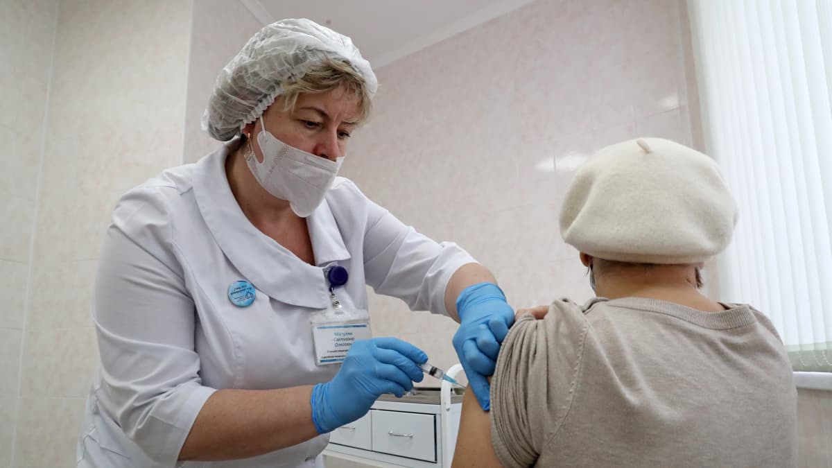 En kvinna i vit basker vaccineras av en kvinna klädd i vit jacka, hårskydd, munskydd samt blå plasthandskar.