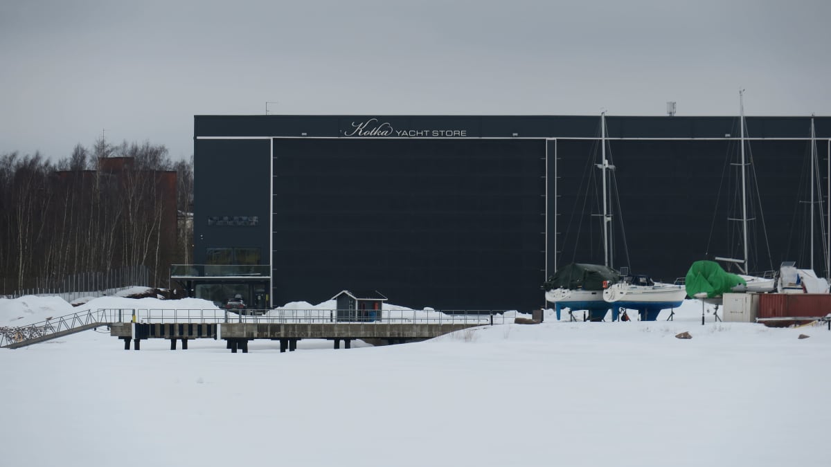Veneiden säilytyshalli Yacht Store Kotkassa ulkoa päin kuvattuna. Tummansininen rakennus, pihalla pieni mökki ja auto.