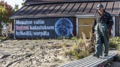 Markku Sotisaari kävelemässä laiturille päin, taustalla on banneri, jossa on hänen kannanosoitus kalastajien hyljeongelmaan.