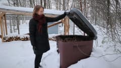 Nainen nostaa kompostin kantaa lumisella pihalla.