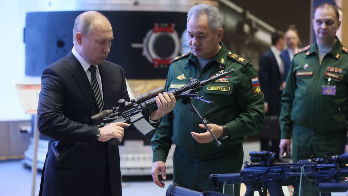 Presidentti Vladimir Putin pitää asetta kädessä ja puolustusministeri Sergei Shoigu seuraa vierestä.