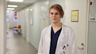 Nainen lääkärin valkoisessa takissa seisoo käytävällä ja katsoo kohti.