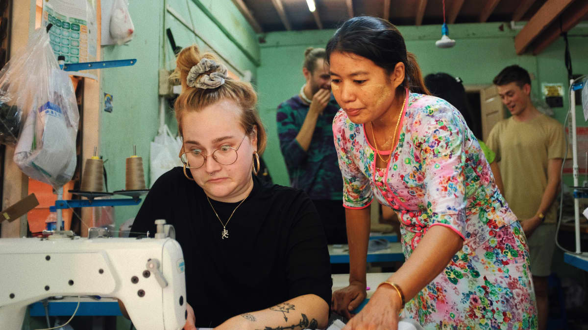 Nuori suomalainen nainen kauhistuneen näköisenä kokeilee ompelukonetta Myanmarissa vaatetahtaalla. Paikallinen nainen opettaa.