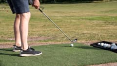 Någon håller på att slå en golfboll med golfklubba. I bilden syns tvp ben, golfboll, golfklubba och golfbanan i bnakgrunden. 
