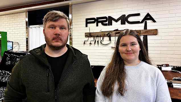 En man och en kvinna i 30-årsåldern står framför en hög bänk, i bakgrunden en tegelvägg med upphängda gamla verktyg och texten Primica.