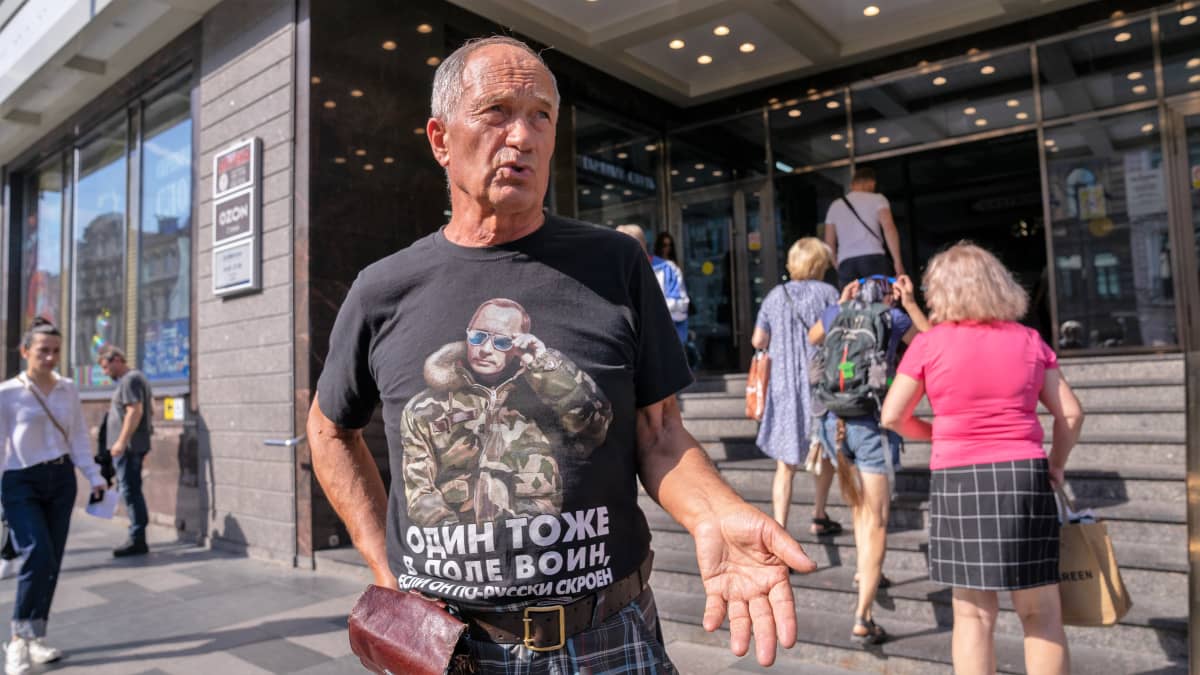 Mies seisoo Suomen viisumikeskuksen rakennuksen edustalla. Hänellä on musta t-paita, jossa on maastoasuisen Putinin kuva.