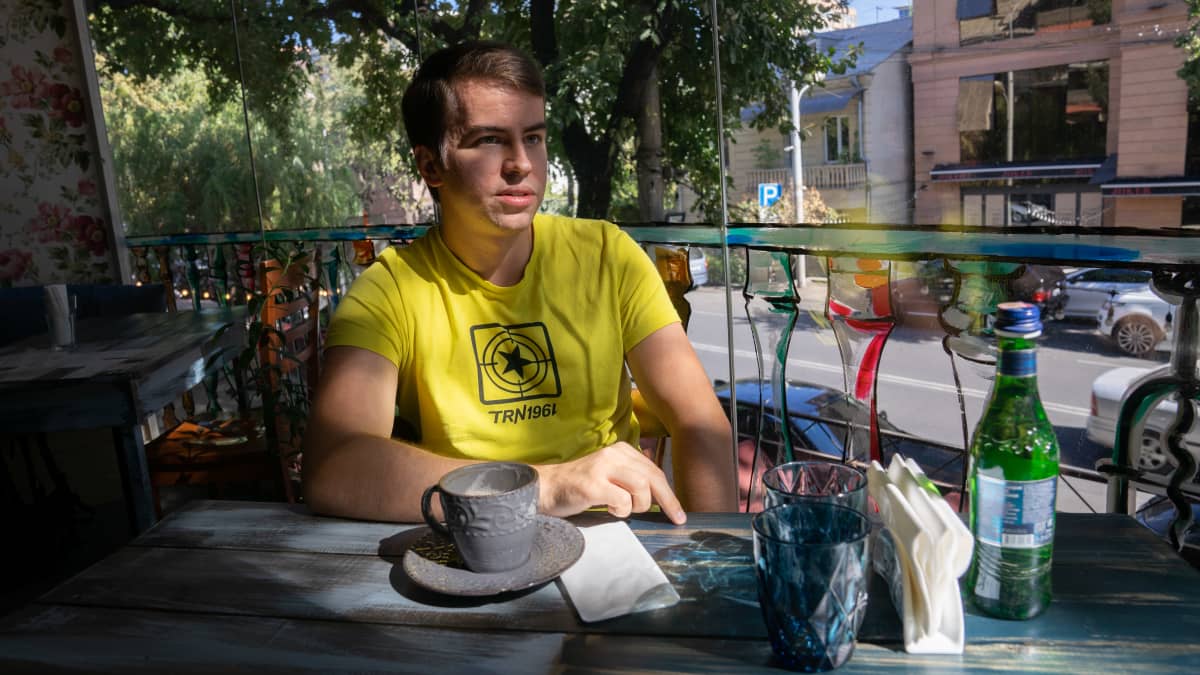 Nuori mies istuu kahvilan pöydän ääressä kahvikuppi ja mineraalivesipullo edessään. Hänellä on keltainen t-paita, jossa on tähden kuva ja teksti TRN1961. Taustalla näkyy aurinkoinen katu.