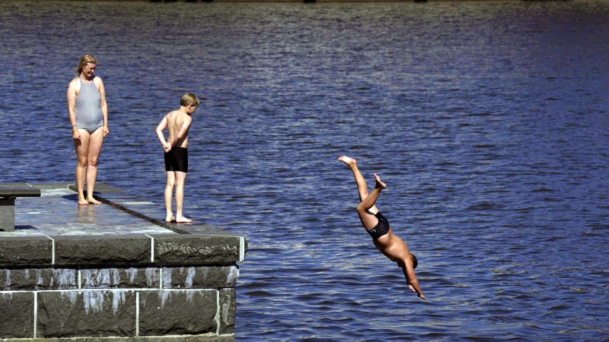 Uimari on hyppäämässä kiviseltä laiturilta kädet ja pää edellä mereen. Laiturilla seisoo kaksi muuta ihmistä, jotka hymyillen katsovat hyppääjää.