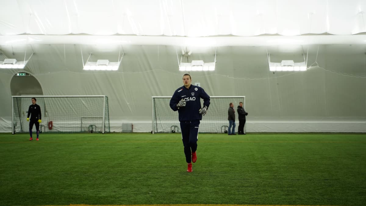 Pelaaja juoksee kohti kameraaa jalkapallohallissa, taustalla tekonurmikentän reunalla kaksi harjoitusmaalia.