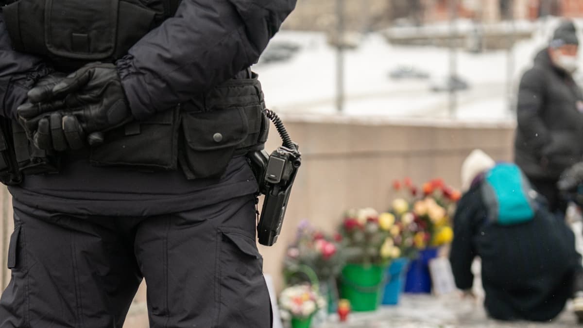 Poliisi seisoo etualalla. Kuvassa näkyy hänen vyöllään oleva pistooli. Kauempana näkyy Nemtsovin muistopaikka, jonne ihmiset tuovat kukkia.
