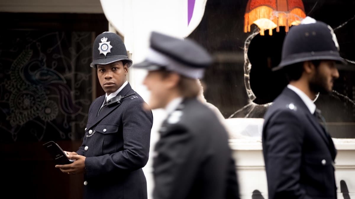 Leroy Logan (John Boyega) bobbyn uniformussa. Taustalla rikottu ikkuna, etualalla muita poliiseja.