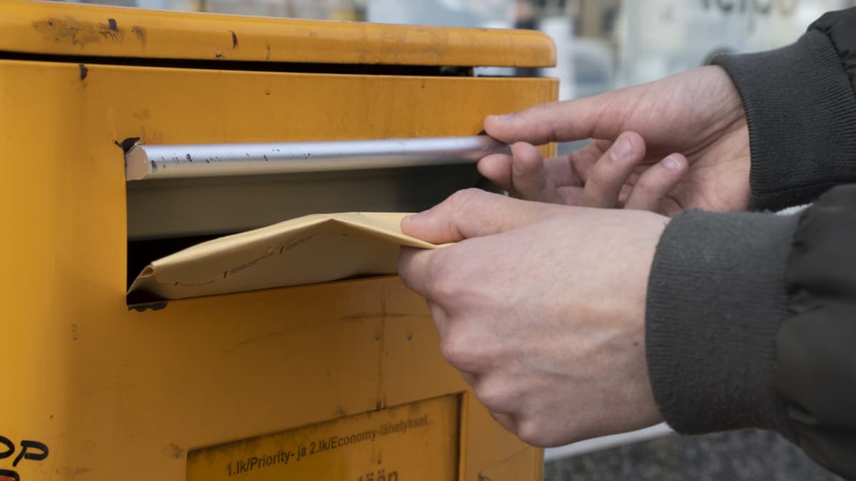 Kädet sujauttamassa ruskeaa kirjekuorta postin oranssiin laatikkoon.