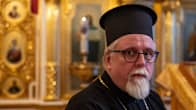 Kuvassa on ortodoksinen pappi seisomassa kirkossa, yllä musta kaapu ja kaulassa koristeellinen risti.