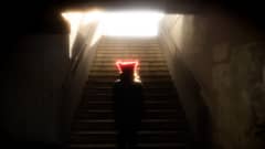 Henkilö punainen pipo päässä auringosta hehkuen pimeiden portaiden alkupäässä. Ylhäällä portaiden päässä aurinko loistaa.