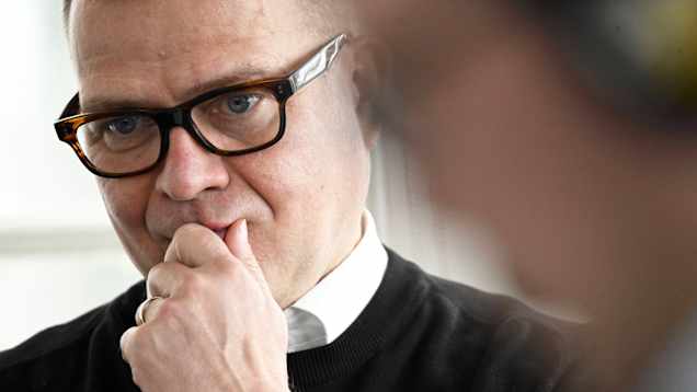 A man with brown glasses, a white shirt and black jumper looks down with a serious expression while holding his chin.