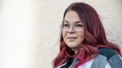 Silmälasipäinen nainen punaiseksi värjätyissä hiuksissa ja ripsipidennyksissä katsoo suoraan kameraan.