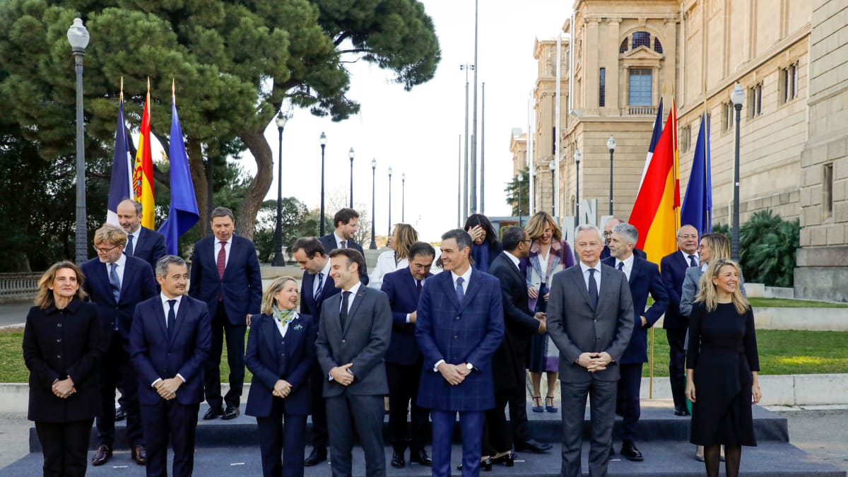 Tummapukuisia miehiä ja naisia juttelee ja naureskelee ryhmäkuvassa kolmella rivillä Espanjan, Ranskan, Katalonian ja EU-lippujen edessä.