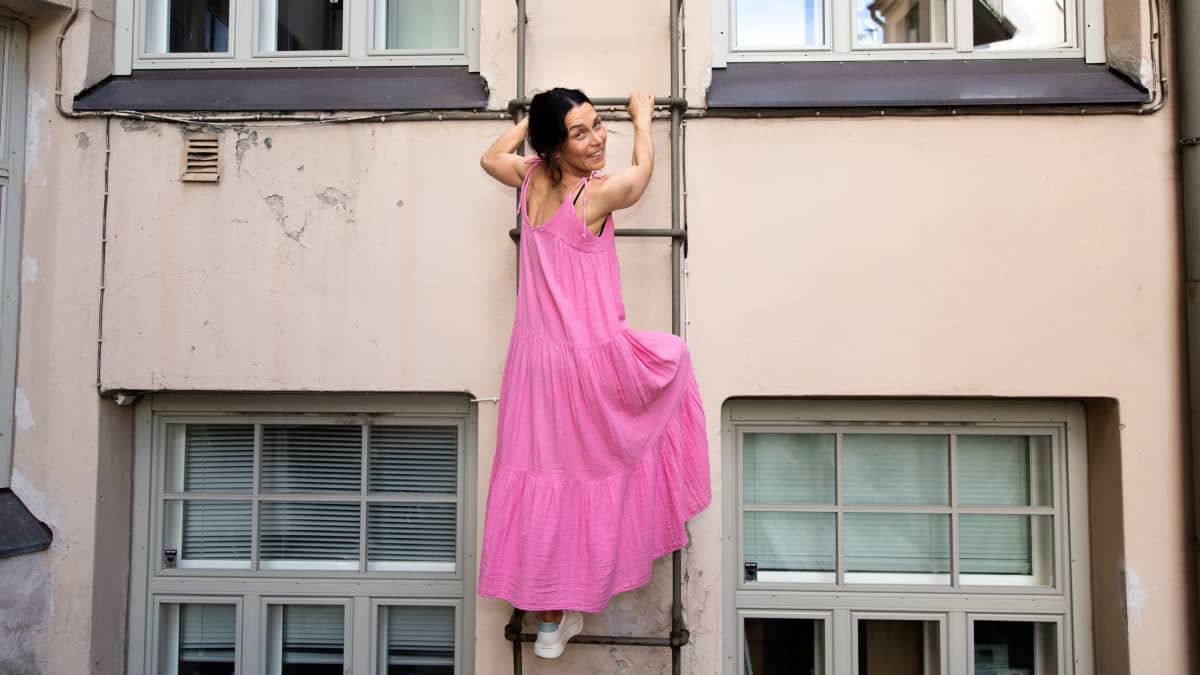 Nainen vaaleanpunaisessa kesämekossa kiipeää tikapuita ylös ja hymyilee. Takana kerrostalon seinä ja ikkunoita. 