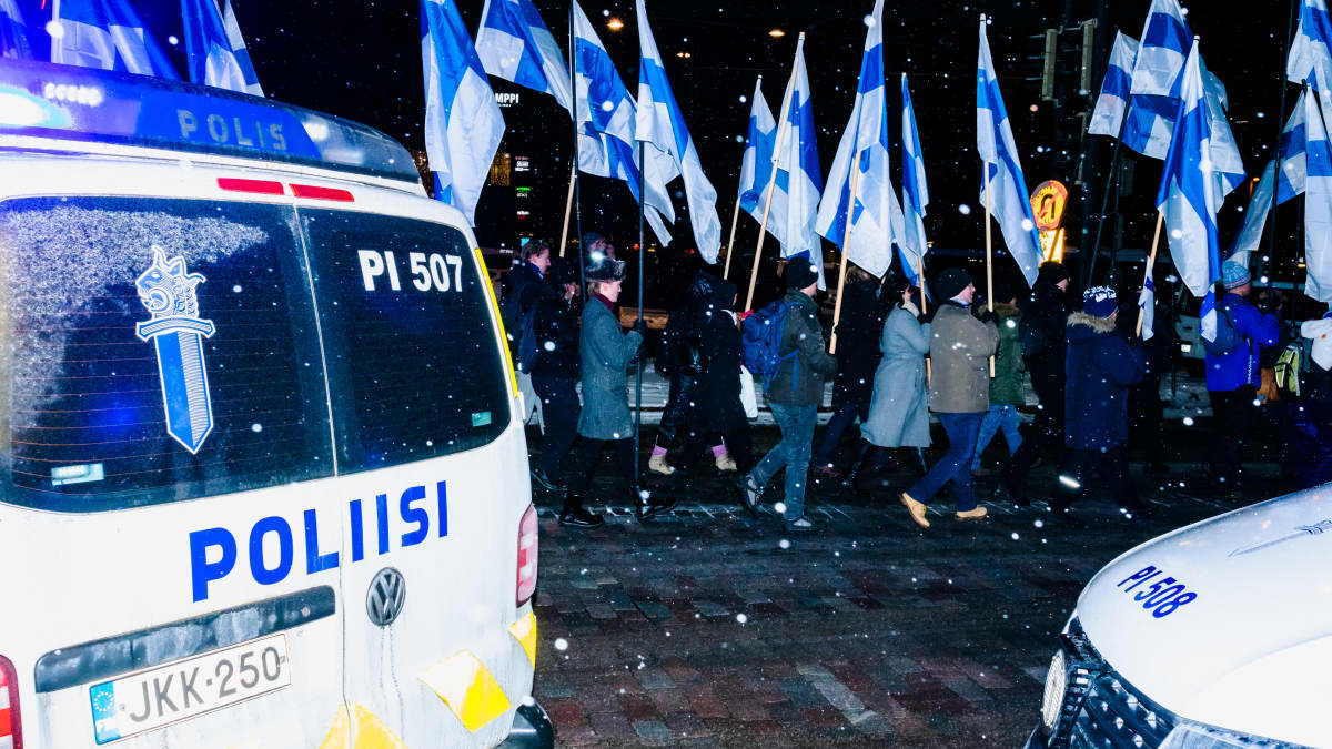 Äärioikeiston mielenosoittajia Suomen lippujen ja iskulauseiden kanssa lumisateessa Mannerheimintiellä poliisiautojen ympäröiminä.