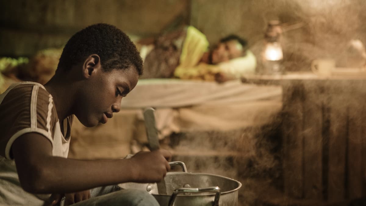 Nuori poika hämmentää jotain kattilassa. Taustalla Nasra (Yasmin Warsame) makaa sängyssä. Kuva elokuvasta Guled & Nasra.