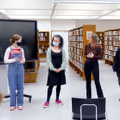 Viisi nuorta naista seisoo rivissä kirjastossa. Keskimmäinen nainen on aikuinen, muut ovat teini-ikäisiä.