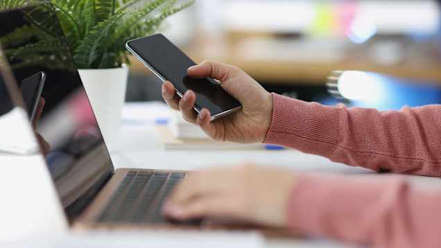 A person's hands seen over a laptop keyboard and holding a smartphone in a brightly lit room.