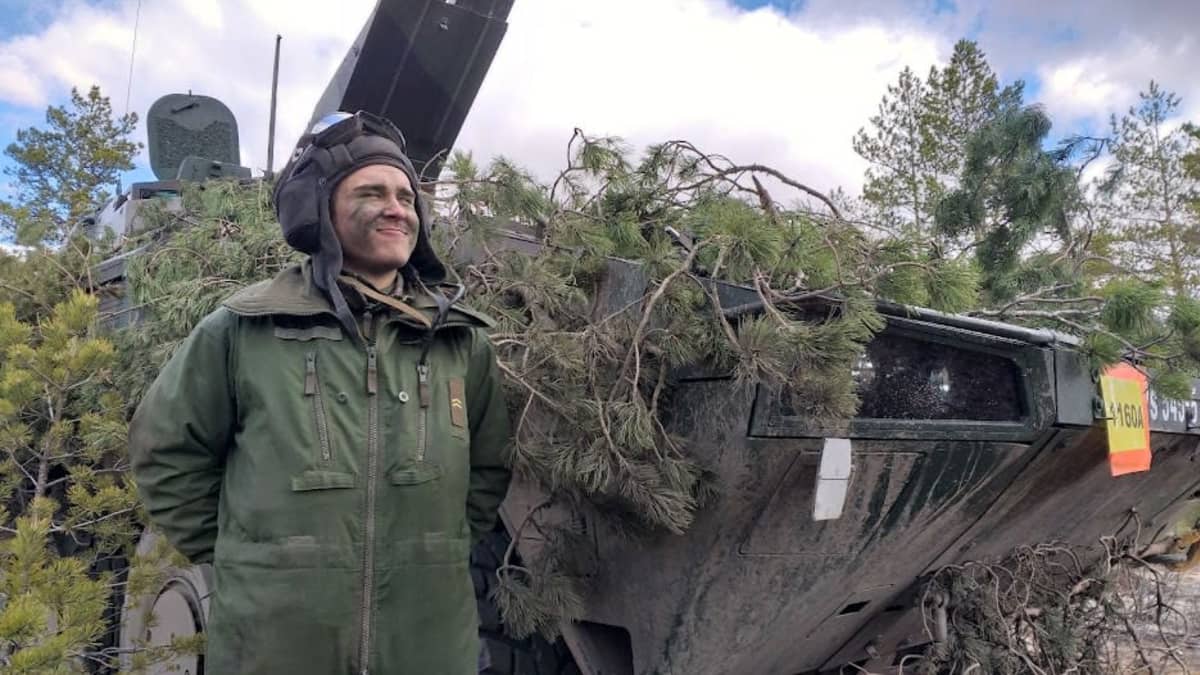 Armeijan haalarissa ja kypärässä oleva nuori mies hymyilee panssarivaunun vieressä.