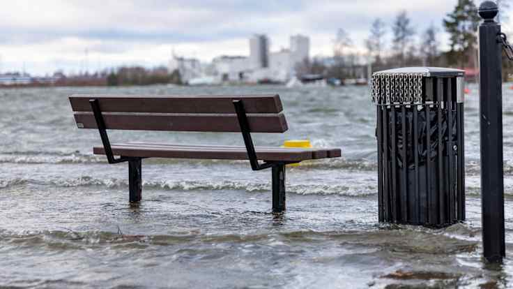 A park bench and a trash can by a shore with elevated water levels.