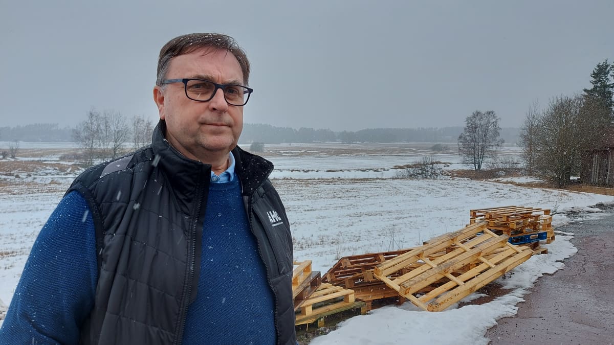 Yrittäjä Pekka Aho seisoo lumisateessa kuormalavojen edessä. Taustalla talvinen maalaismaisema.