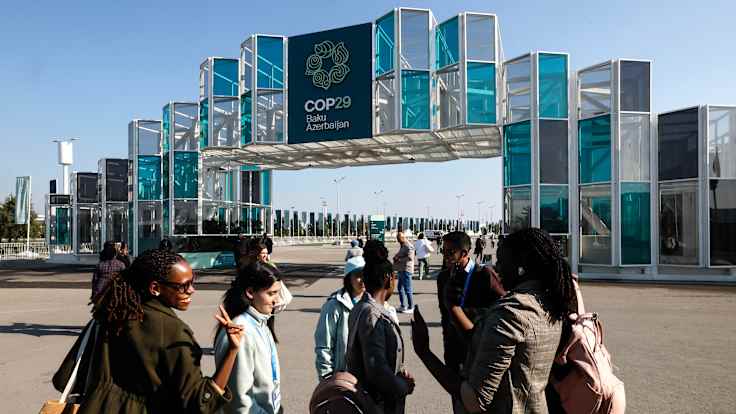 A group of young people stand talking in front of a glass-and-metal gateway reading COP29 Baku Azerbaijan.