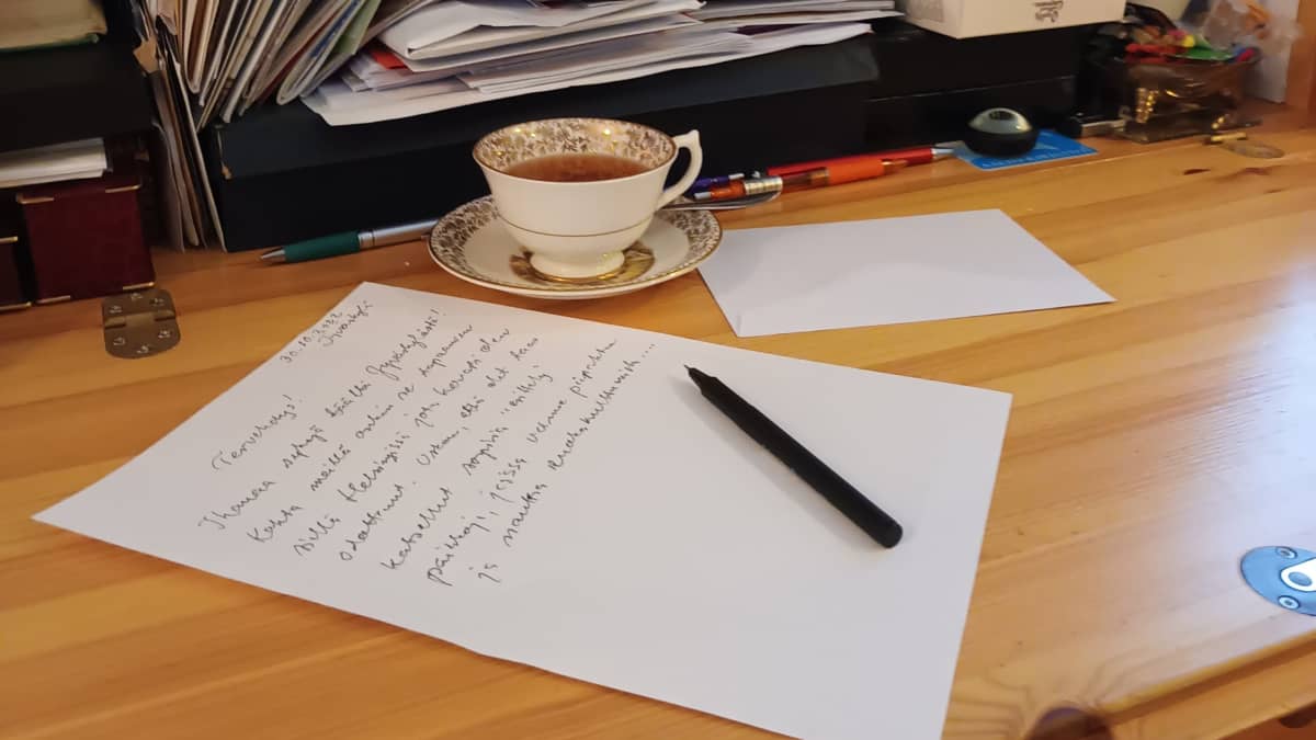 Pöydällä on kirje, jota kirjoitetaan parhaillaan. Vieressä on teekuppi ja kirjekuori. 