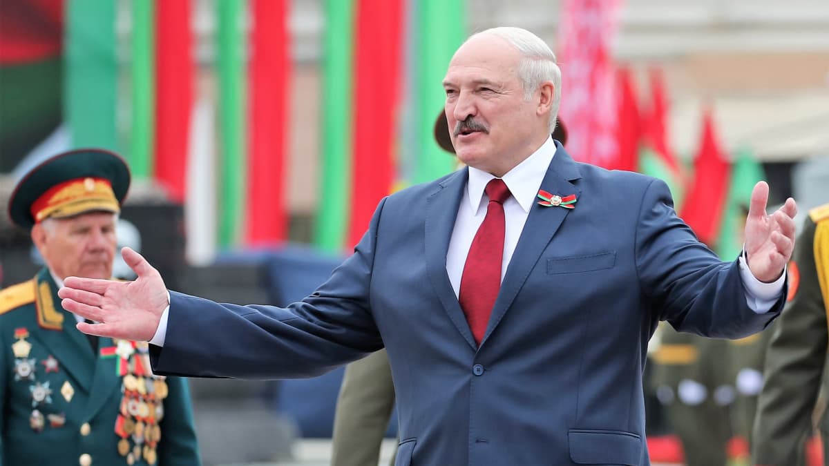 Lukashenka levittää käsiään hymyillen. Hänellä on tumma puku, punainen kravatti. Taustalla seisoo vanha mies univormussa.