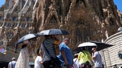 Ihmisiä Sagrada Familian edessä kuumana kesäpäivänä sateenvarjot auringon suojana.