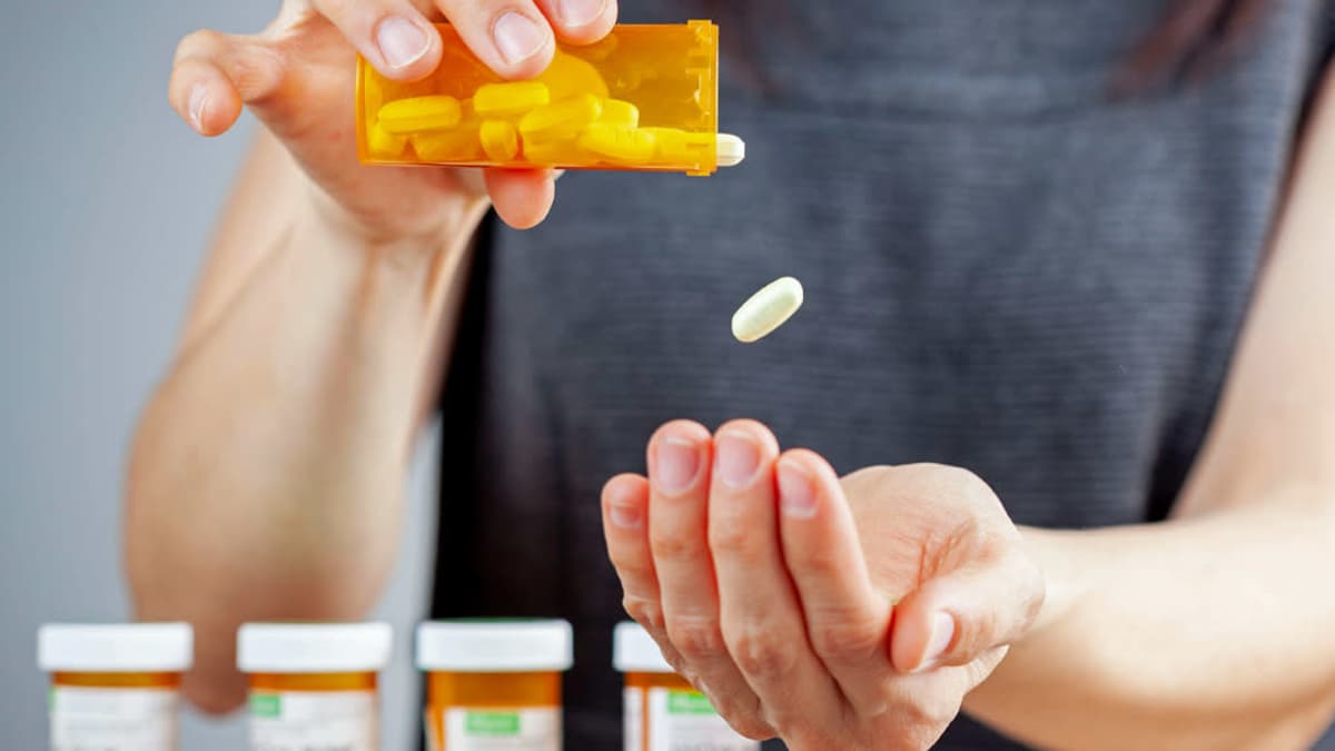 Nainen kaataa tabletteja kämmenelleen, edessä pöydällä on rivi muita pilleripurkkeja. 