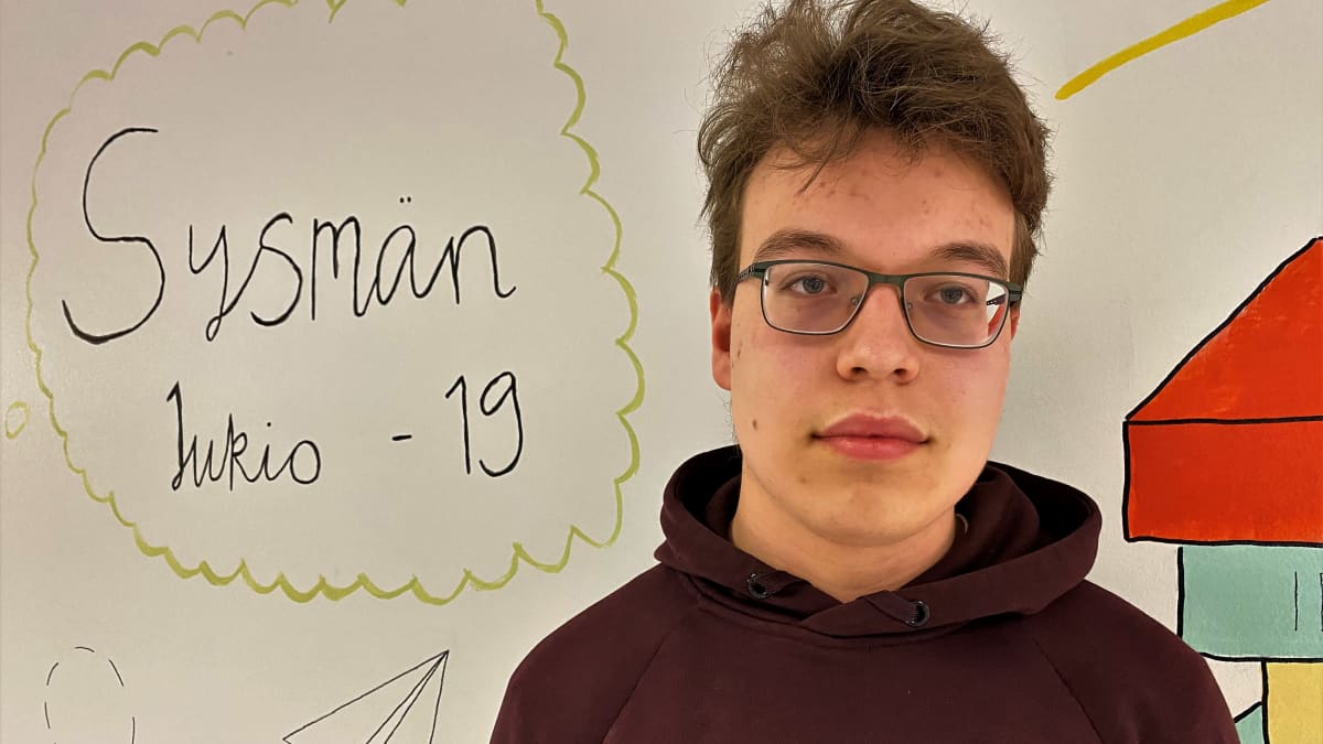 Lukiolainen Lauri Hildén ruskeassa hupparissa seisoo piirroksen edessä, jossa teksti: Sysmän lukio -19. 