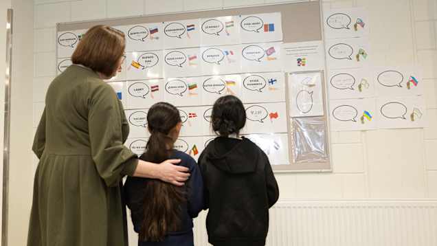 A woman and two girls seen from behind, standing looking at a bulletin board with signs with flags from all over the world.