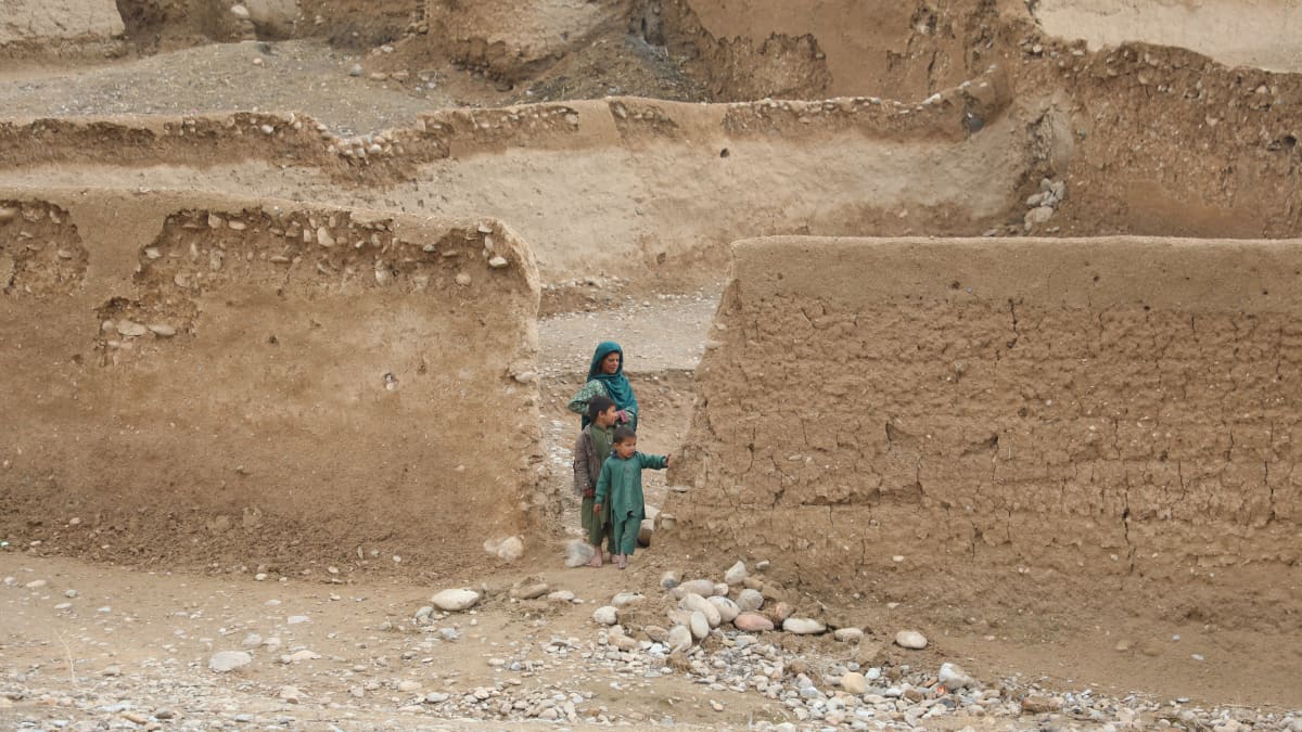 Nainen ja kaksi lasta seisoo savitiilistä rakennetun aidan aukossa.