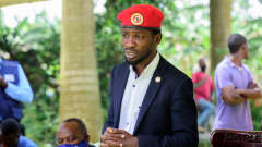 Bobi Wine puhuu tiedotustilaisuudessa ulkotilassa. Hänellä on tumma puku, vaalea kauluspaita ja päässään punainen baretti.