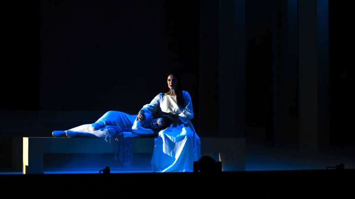 Tummatukkainen nainen istuu ja laulaa pimeällä näyttämöllä toinen nainen makaa penkillä pää laulavan naisen sylissä.