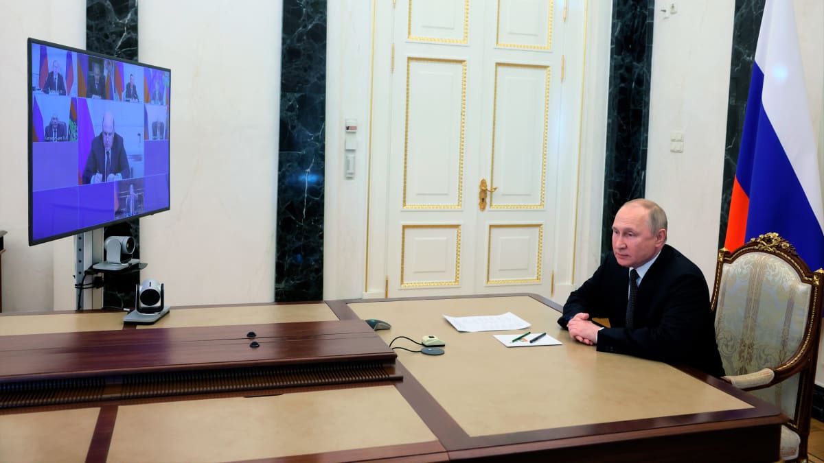 Putin istuu kuvan oikeassa laidassa pöydän päässä kädet ristissä edessään. Takana näkyy Venäjän lippu. Pöydällä on televisioruutu, jossa näkyy turvallisuusneuvoston jäseniä.