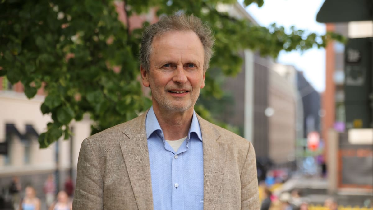 Professori Kimmo Nuotio Helsingin Yliopiston edessä. Taustalla ihmisvilinää ja puun oksia vihreine lehtineen. Kimmolla on päällään ruskeanharmaa pikkutakki ja sininen kauluspaita.