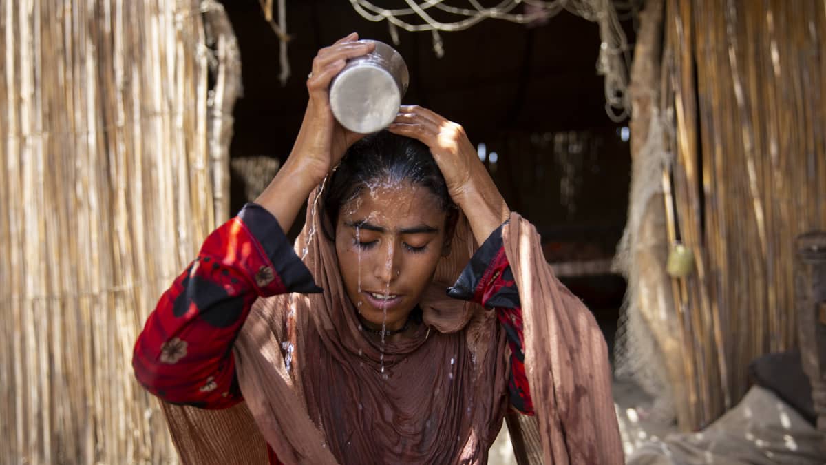 Nainen kaataa kannusta vettä päälleen kaislamökin edustalla silmät suljettuina.