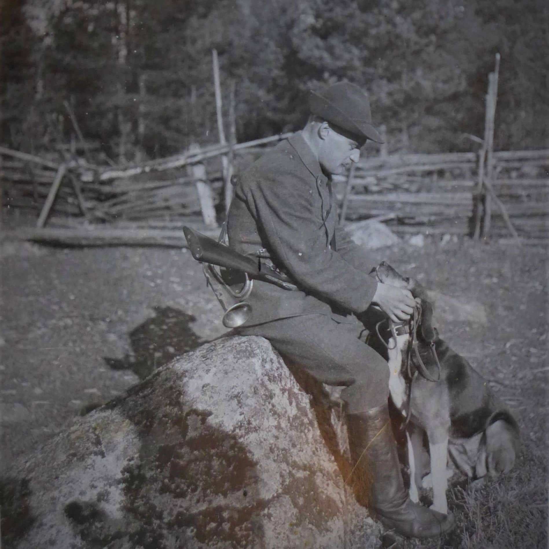 Mies (metsänhoitaja Torsten Rosenbröijer) istuu kivellä haulikko selässä ja rapsuttaa ajokoiraa.