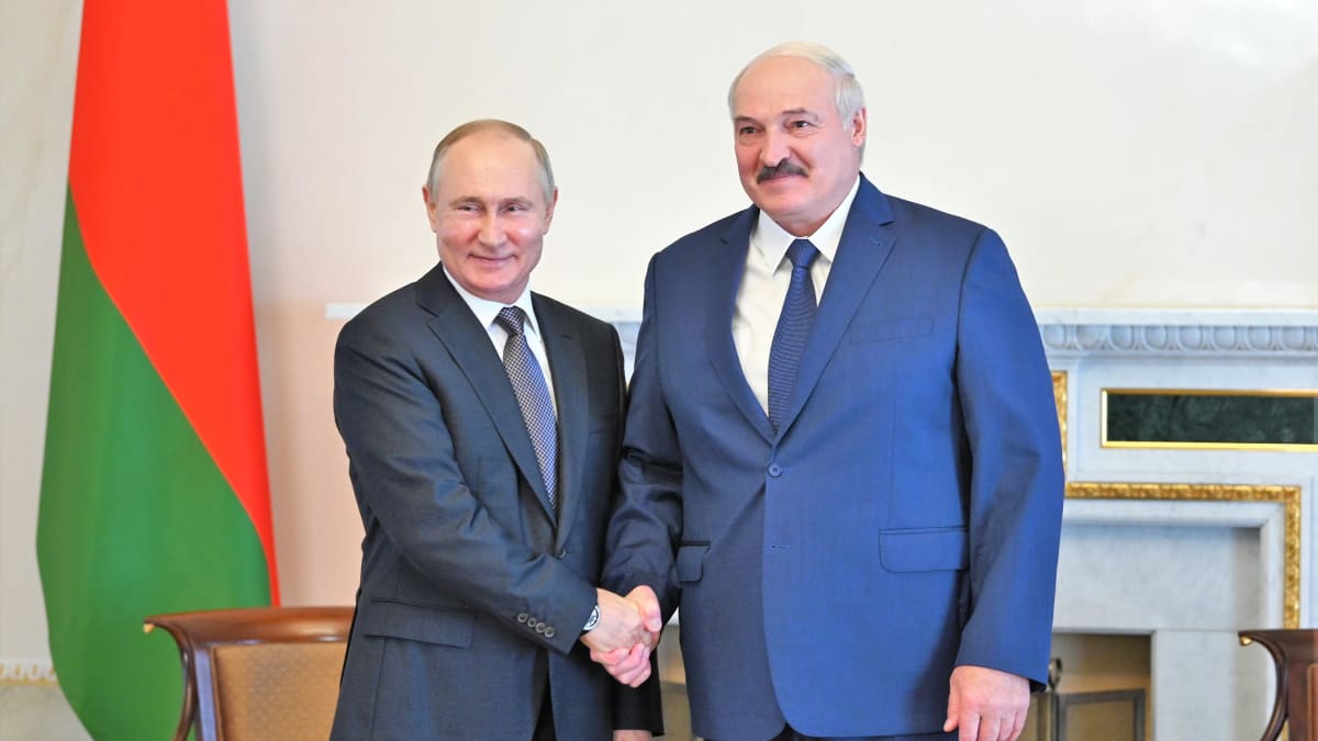 Putin kättelee Lukašenkaa ja hymyilee kameralle. Myös Lukašenka hymyilee. Miehet poseeraavat kameroilla timmissa puvuissa. Taustalla huoneessa näkyy Valko-Venäjän lippu.