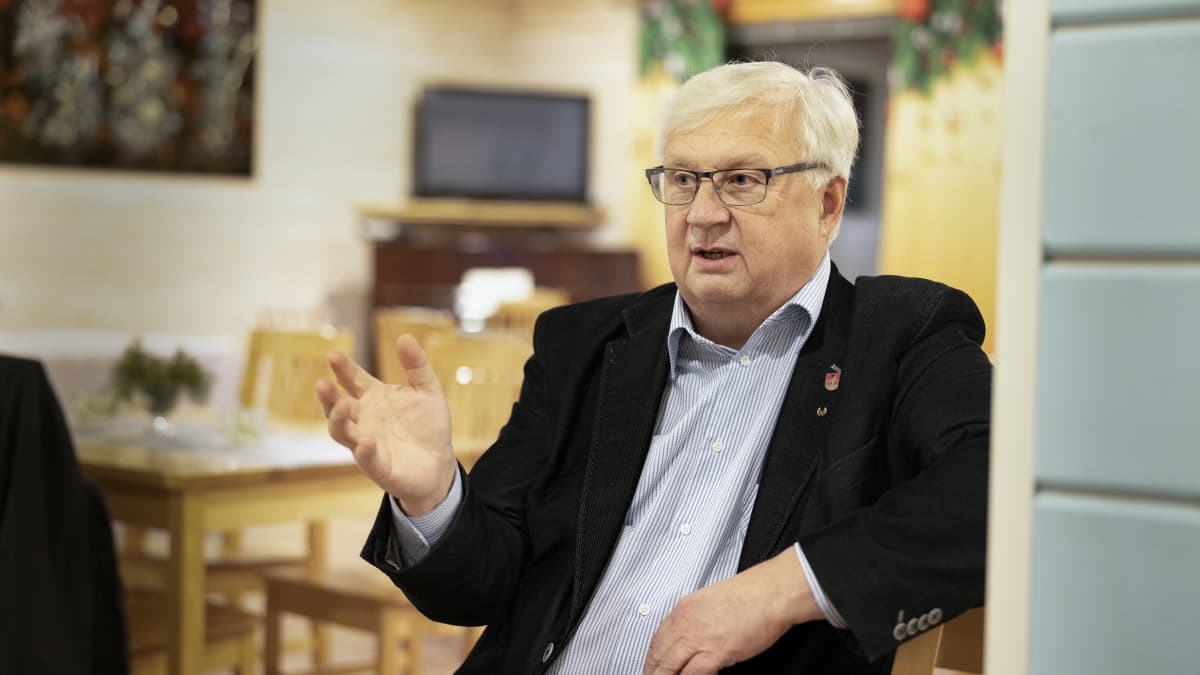 Pertti Huurtola istuu Karjalaisen kotileipomon juhlatilassa ja puhuu käsi heiluen.