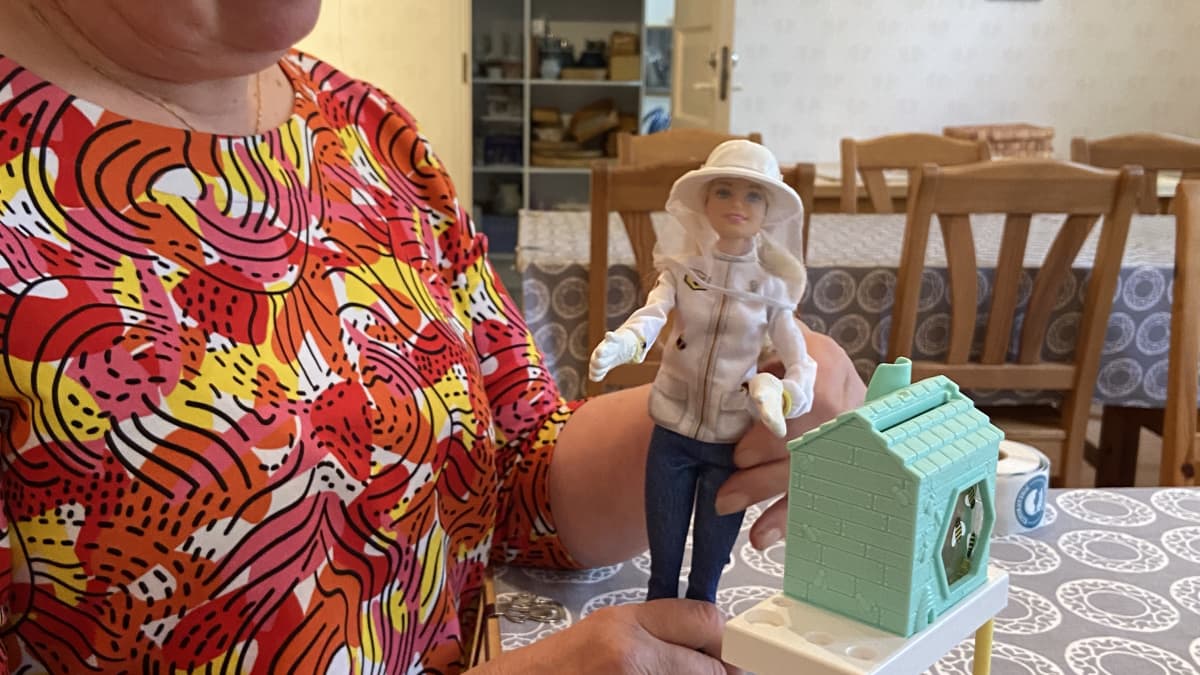 Luomumarjatilan emäntä Seija Halme esittelee pöydällä seisovaa mehiläishoitaja-barbie-nukkea. Nuken edessä on tilan mehiläisten hunajaa. Hunajapurkin kannessa on uusi lähiruuan  alkuperämerkintä " P