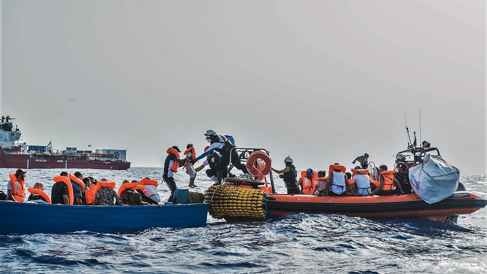 Siirtolaisia autetaan sinisestä veneestä oranssireunaiseen kumiveneeseen. Sukelluspukuinen mies ottaa juuri vastaan lasta. Taustalla näkyy punavalkoinen Ocean Viking -alus.