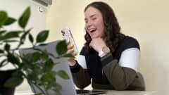 Mirka Helander istuu pöydän ja kannettavan tietokoneen äärellä ja selaa puhelinta naureskellen