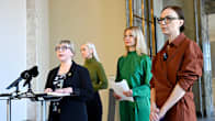 Four women stand in a corridor holding papers, one speaking at a lectern.
